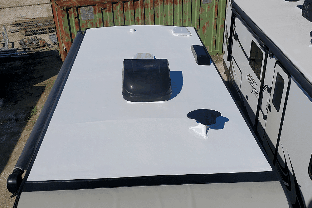 flexarmor installation, RV repair in Laplace, LA at RV Masters. Top view of a white RV roof with various fixtures, including an air conditioning unit, vent covers, and an antenna. The RV is parked beside another trailer, with a rusty shipping container and fencing in the background.