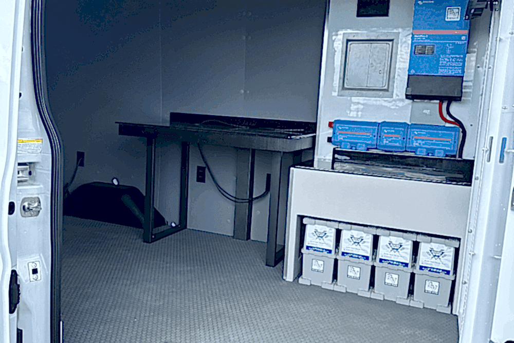 Nick Underhill's custom van build, RV repair in Laplace, LA at RV Masters. Electrical system setup in the back of a service van, featuring mounted batteries, inverter, and power control units with a small workbench and organized wiring.