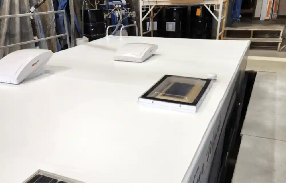 FlexArmor Roof Coating, RV repair in Laplace, LA at RV Masters. A white, freshly coated RV or trailer roof inside a workshop, featuring two low-profile white rooftop units and a rectangular roof vent with a transparent cover. Industrial shelves, tools, and large black barrels are visible in the background.